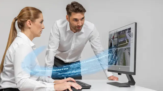 Frau sitzt vor Überwachungsbildschirm, Mann beugt sich über den Tisch, um mit reinzusehen