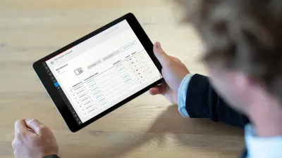 Person hält Tablet mit Fire System Analyzer-Anwendung, die ein Betriebsbuch mit chronologisch aufgelisteten Ereignissen und Alarmzählern vom Juni und Juli 2024 anzeigt. Die Tabelle enthält Datumsangaben, Statuswerte und verschiedene Ereignissymbole.