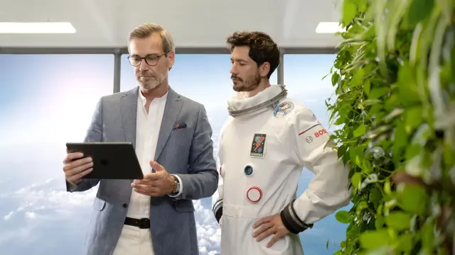 Ein Geschäftsmann schaut gemeinsam mit einem Astronauten auf ein Tablet. Sie stehen vor einem Fenster mit Wolken im Hintergrund.