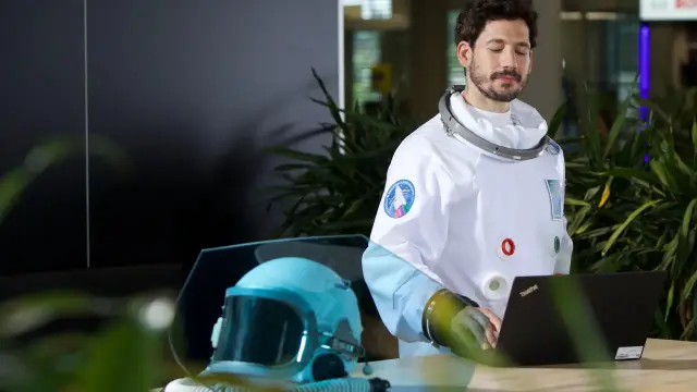 Mann in weißem Raumanzug tippt auf einem Laptop. Im Vordergrund steht sein Helm auf einem Tisch, überdeckt von einem halbtransparenten, blauen Schutzschild.
