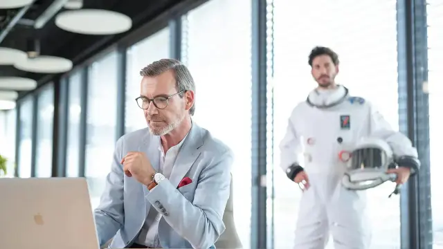 Ein Mann im Business-Outfit arbeitet konzentriert an seinem Laptop, während im Hintergrund ein Astronaut als symbolischer Beschützer steht. 