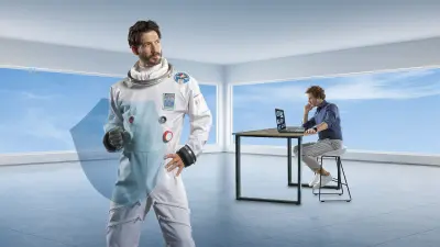 Mann im weißen Raumanzug hält ein transparentes blaues Schutzschild, im Hintergrund arbeitet ein Mann in einem modernen Büro am Laptop .