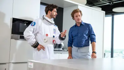 NEXOSPACE Datanaut holds a black tablet and points to it; on the right stands a man with curly brown hair in a blue shirt, gesturing in a modern office environment.