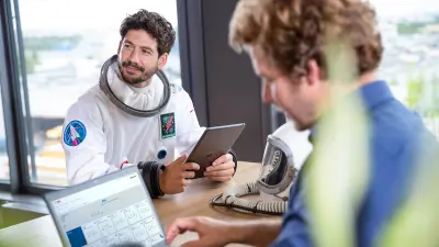 NEXOSPACE Datanaut holds a black tablet and sits at a table opposite a man with brown hair in a blue shirt who is working on a laptop, modern office environment with large windows