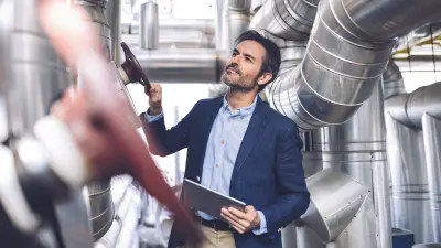 Man in dark business suit and light blue shirt holding black tablet and looking up, standing in industrial setting with silver pipes and ventilation systems on the ceiling, blurred purple object in foreground