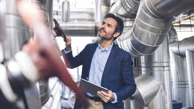 Man in dark business suit and light blue shirt holding black tablet and looking up, standing in industrial setting with silver pipes and ventilation systems on the ceiling, blurred purple object in foreground