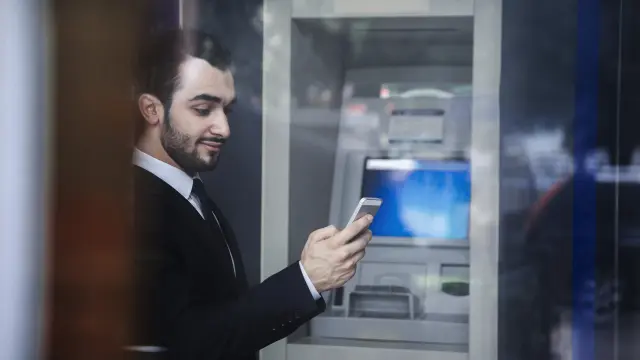 Dunkelhaariger Mann mit Bart steht vor einem Geldautomaten mit Smartphone in der Hand.