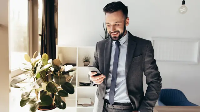 Bärtiger Mann im Anzug sieht auf sein Smartphone und lacht.
