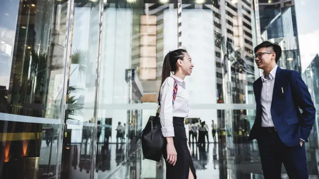 Mann und Frau in Business Kleidung stehen von einem Eingang eines gläsernen Gebäudes.