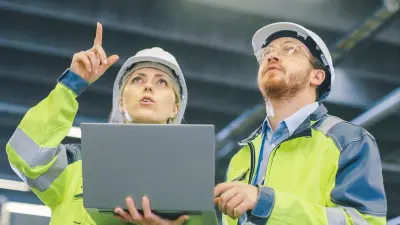Frau mit Laptop spricht mit einem Kollegen, beide tragen Warnweste und Bauhelm