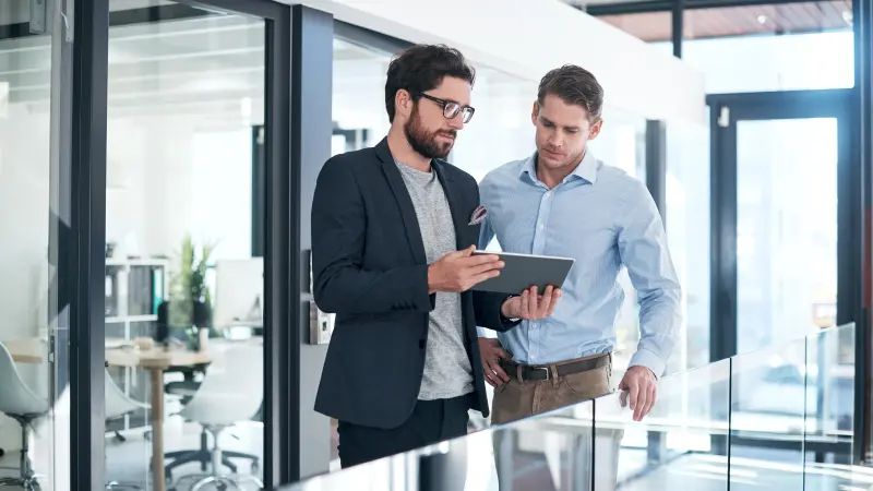 Zwei Geschäftsmänner mit Tablet besprechen sich in einem modernen Bürogebäude miteinander