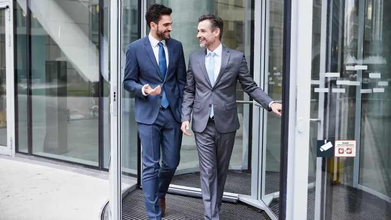 Zwei Bankmitarbeiter im Anzug verlassen das Bankgebäude durch eine Drehtür