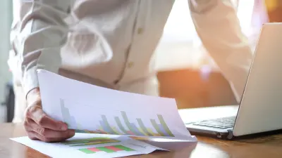 Person im Büro beugt sich über einen Tisch und analysiert ausgedruckte Diagramme und Grafiken neben einem Laptop.