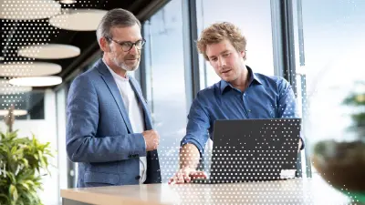 Zwei Männer stehen vor einem Tisch und schauen auf einen Laptop. 
