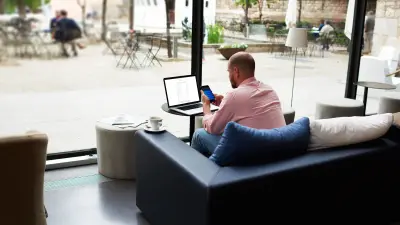 Mann sitzt in der Lobby eines Hotels vor einem Laptop und hat Mobiltelefon in der Hand.