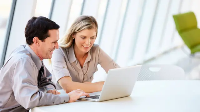 Dunkelhaariger Mann und blonde Frau sitzen am Tisch und sehen gemeinsam in ein Laptop.