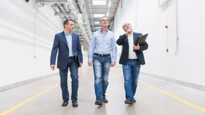 Drei Männer in Hemd und Sakko gehen durch eine helle, moderne Industriehalle und besichtigen die Anlage. Einer der Männer deutet nach oben.