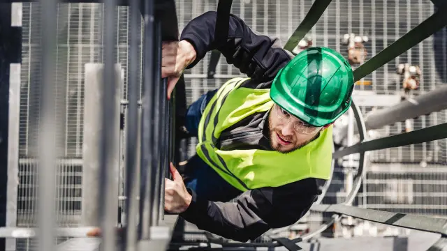 Aus der Vogelperspektive wird ein Monteur mit grünem Schutzhelm und leuchtend gelber Warnweste gezeigt, der auf einer Metallstruktur einer Industrieanlage klettert.