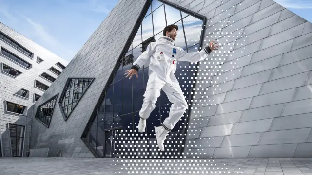 Ein Mann in einem weißen Astronautenanzug schwebt vor einem modernen, architektonisch auffälligen Gebäude mit Metallfassade. Eine digitale Grafik aus weißen Punkten umgibt ihn.