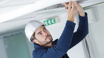 Ein Techniker mit weißem Schutzhelm und blauem Arbeitskittel führt konzentriert Wartungsarbeiten an einer Deckenleuchte oder einem Kabelkanal durch.
