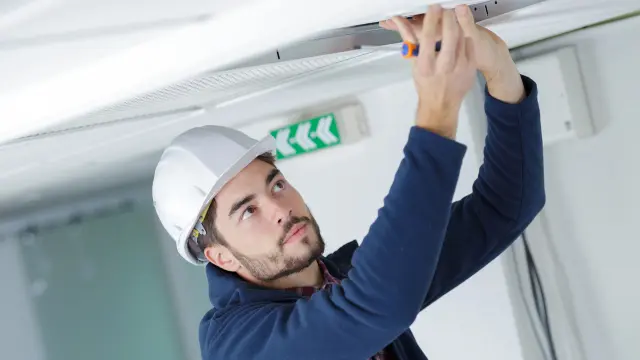 Ein Techniker mit weißem Schutzhelm und blauem Arbeitskittel führt konzentriert Wartungsarbeiten an einer Deckenleuchte oder einem Kabelkanal durch.