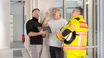 3 Männer, inklusive Feuerwehrmann, stehen zusammen mit Tablet und schauen zu einem Brandmelder hoch.