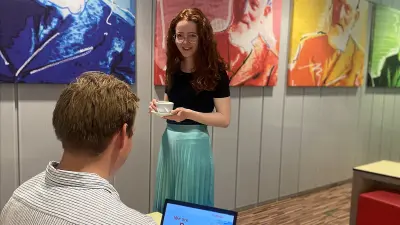 Romina läuft mit einer Kaffeetasse in der Hand in der Cafeteria zu einem Kollegen, der an einem Tisch sitzt
