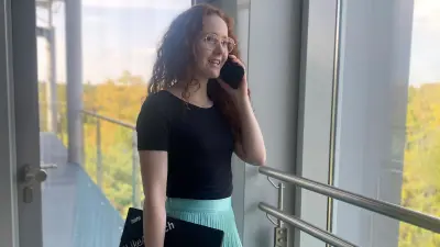 Romina steht an einem großen Fenster mit Blick auf einen Wald und telefoniert mit ihrem Handy