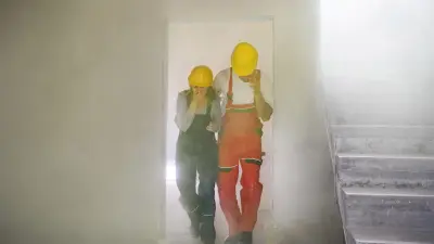 Man and woman in boilersuits wearing yellow helmets covering their noses and escaping from a smoke filled staircase