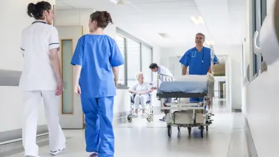 A hospital corridor with several staff walking along it, one carrying a stretcher and another assisting a lady in a wheelchair.