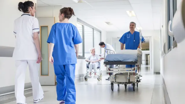 A hospital corridor with several staff walking along it, one carrying a stretcher and another assisting a lady in a wheelchair.